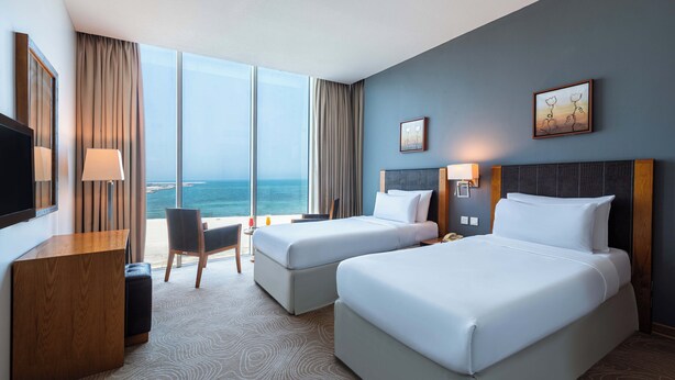 A hotel room with two beds, a desk, a chair, and a large window offering a view of the ocean. - Razana Al Jubail (Al Jubail)