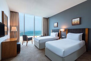 Habitación de hotel con dos camas, un escritorio, una silla y un amplio ventanal con vista al océano.