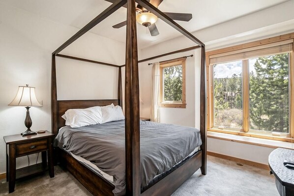 Una cama con dosel, un ventilador de techo, una mesita de noche con lámpara y una ventana con vistas a árboles.
