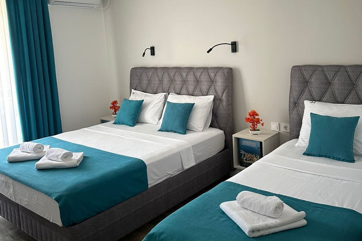 A hotel room with two beds, each with a grey headboard, white bedding, and teal throw pillows. There is a small table with a lamp, a wall-mounted air conditioner, and a window with blue curtains.