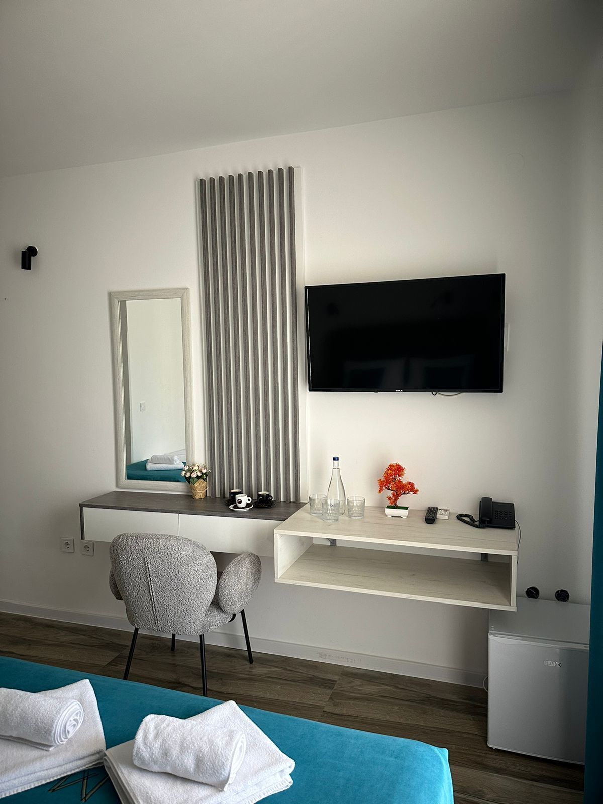 A hotel room with a bed, a desk with a chair, a TV, a mirror, a radiator, a small fridge, and a window with blinds.
