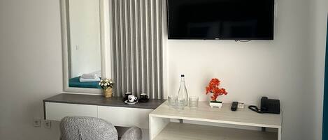 A hotel room with a bed, a desk with a chair, a TV, a mirror, a radiator, a small fridge, and a window with blinds.