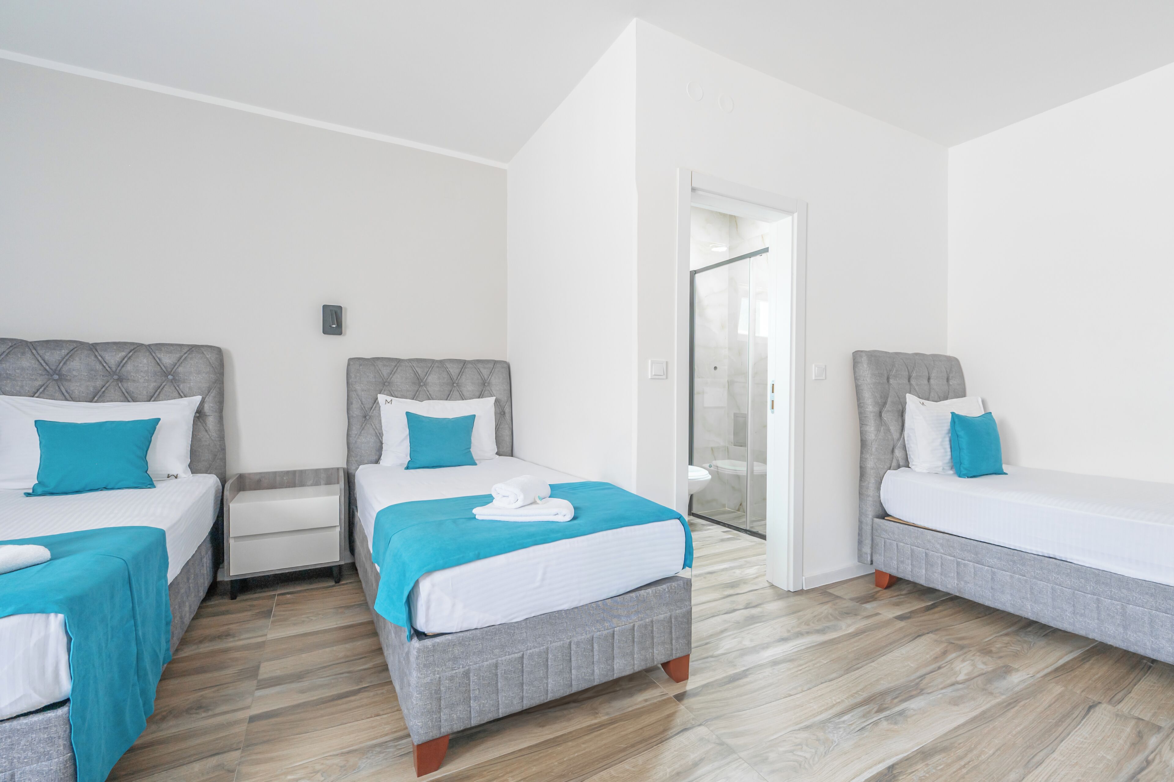 A hotel room with three beds, a bathroom with a glass door, and a wooden floor.