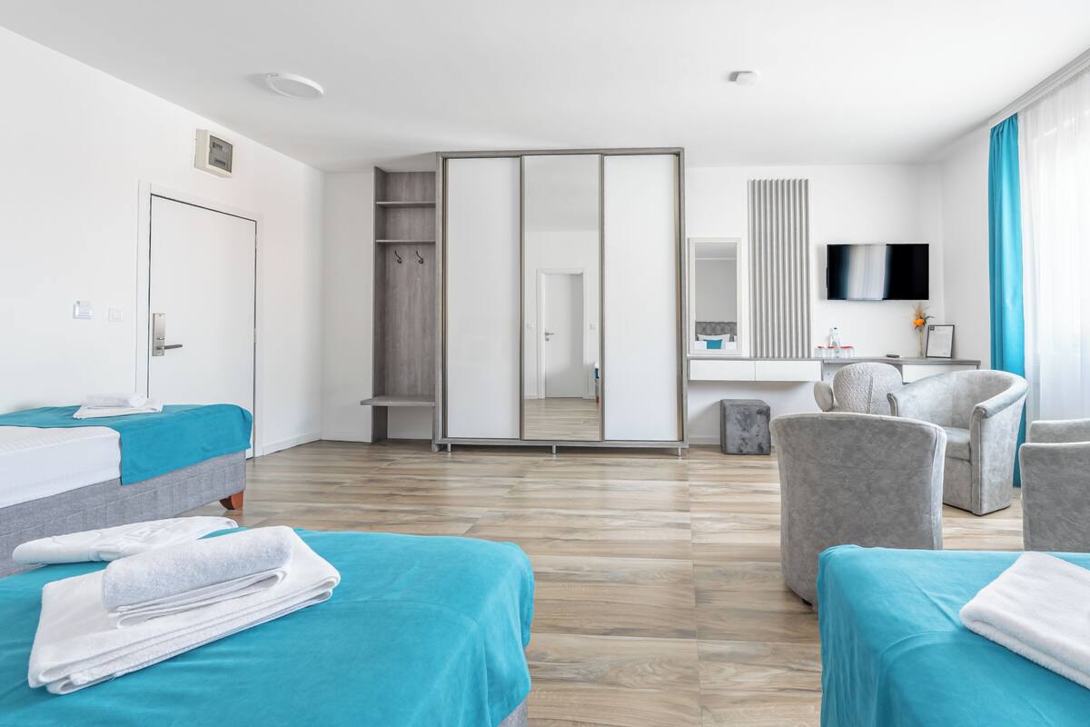 A hotel room with a bed, two bedside tables, a wardrobe, a seating area with a chair, a TV, and a bathroom with a mirror and sink.