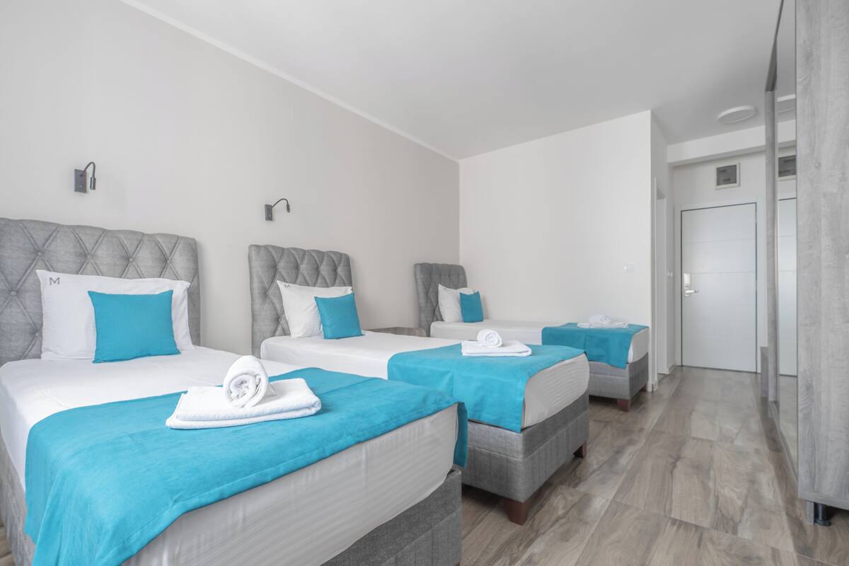 A hotel room with three beds, each with a grey headboard, white bedding, and blue accents.