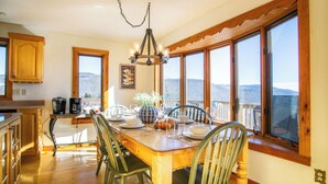 A dining area with a wooden table and chairs, a chandelier, and large windows offering a mountain view.