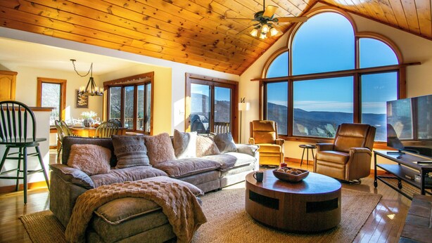 A cozy living room with a wooden ceiling, large windows offering a mountain view, a fireplace, and comfortable seating.