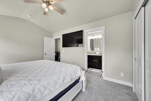 Un dormitorio con cama, ventilador de techo, televisor, baño con espejo y puerta que da a otra habitación.