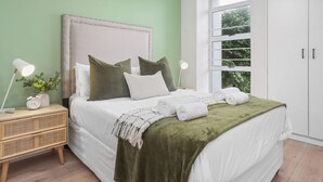 A neatly made bed with a green throw blanket, white bedding, and two rolled towels. A bedside table with a lamp and a clock is visible. - The Sands BG3 - Two Bedroom Apartment, Sleeps 4 (Cape Town)