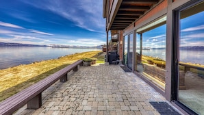 Un patio frente al agua con un banco de madera, una parrilla y vistas a un lago y montañas.