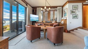 Amplia sala de estar con un gran ventanal corredizo que da a un balcón, comedor con mesa de madera y sillas de cuero, y televisor montado en la pared.