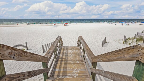 Eine Holztreppe führt zu einem Sandstrand mit Sonnenschirmen und Badegästen.