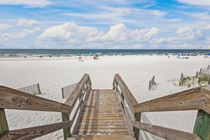 Un escalier en bois mène à une plage de sable où l’on trouve des parasols et des personnes qui se prélassent au soleil.