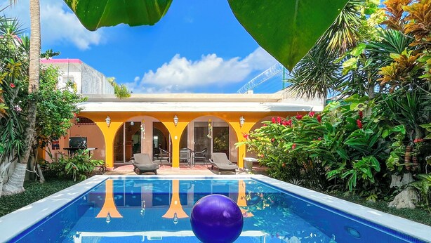A swimming pool with a clear blue water surface, surrounded by tropical plants and a yellow building with outdoor seating.