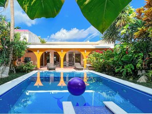 Una piscina con superficie de agua azul cristalina, rodeada de plantas tropicales y un edificio amarillo con mesas al aire libre.