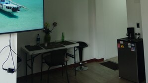 A room with a flat-screen TV displaying a tropical scene, a small dining table with chairs, a refrigerator, and a coffee maker.