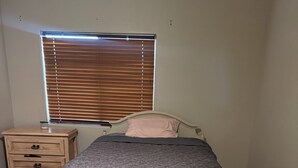 Ein Schlafzimmer mit einem Bett, einem Nachttisch, einem Fenster mit Jalousien und einem Fliesenboden.
