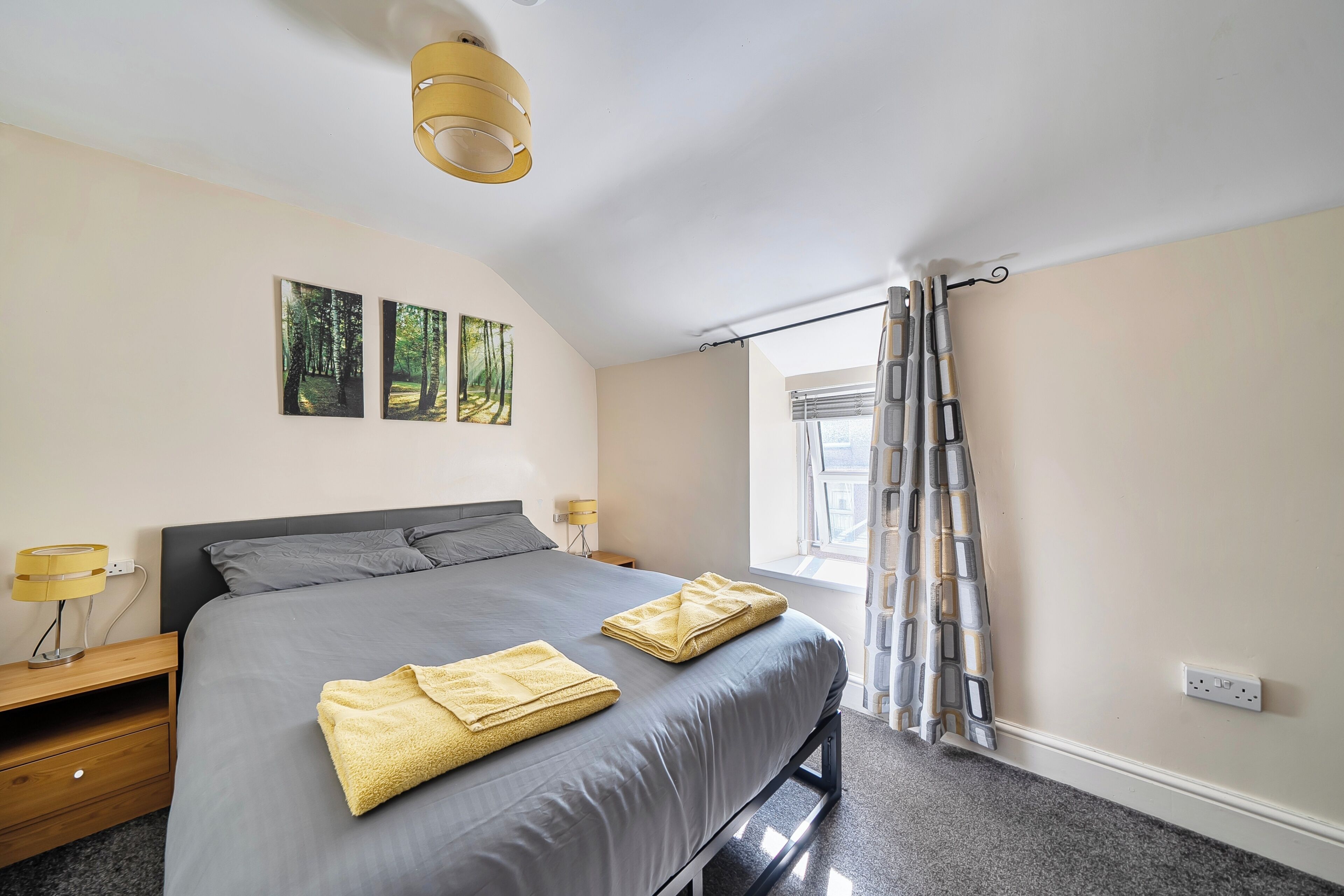 A bedroom with a bed, two bedside lamps, a window with curtains, and artwork on the wall.
