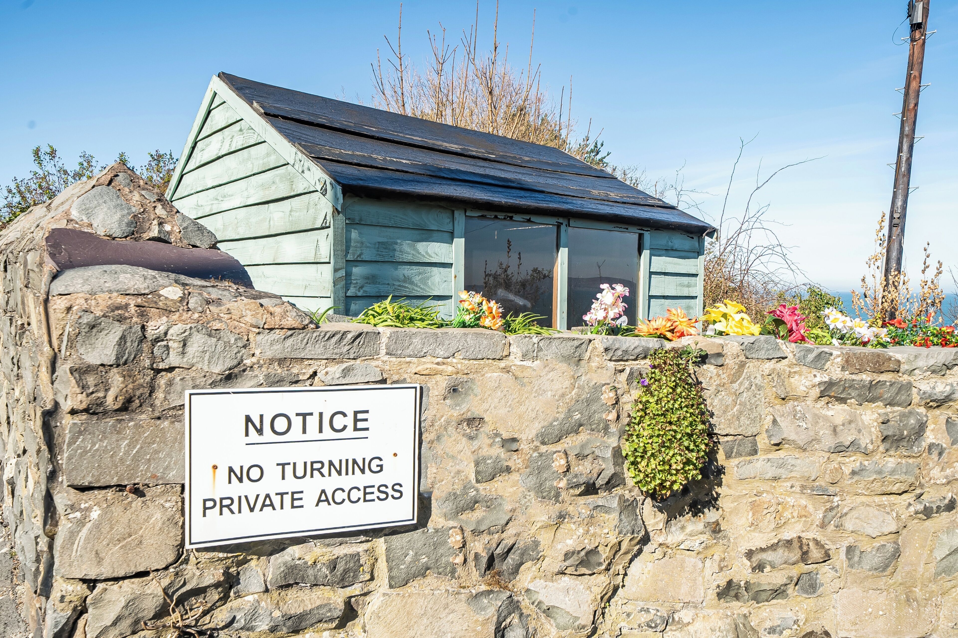 A small, rustic building with a sloped roof, a stone wall with a 'NO TURNING PRIVATE ACCESS' sign, and a variety of colorful flowers in front.