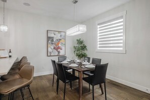 A dining area with a wooden table set for four, black chairs, a chandelier, a potted plant, and a large window with blinds.