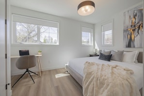 A bedroom with a bed, a desk, and a chair. There are windows and a painting on the wall.