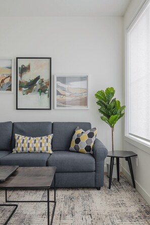 A modern living room with a grey sofa, two patterned cushions, a coffee table, and a potted plant.