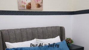 Een bed met witte beddengoed en een blauw kussen, een klein nachtkastje met een plantje, en een aan de muur hangend schilderij.