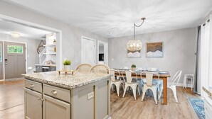 Tilava keittiö, jossa on keskellä sijaitseva saareke, puulattia sekä ruokailutila, jossa on valkoiset tuolit ja koristeellinen kattokruunu.