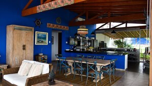 Een ruime woonkamer met een eettafel en stoelen, een rieten bank en een keuken met een bar.