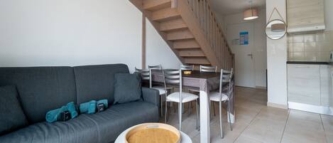 A modern living room with a grey sofa, a dining table with chairs, a wooden staircase, and a kitchen area with white cabinets.