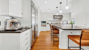 A modern kitchen with white cabinetry, a stainless steel refrigerator, a built-in oven, and a marble backsplash. There are two bar stools and pendant lights above the island. - 5BDRM Renovated Historic | Downtown| Shuttle| Bike Path (Stowe)