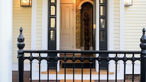 A front porch with a black wrought iron railing, a wooden door with glass panels, and a decorative wall with a patterned design. - 5BDRM Renovated Historic | Downtown| Shuttle| Bike Path (Stowe)