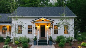 A white house with a front porch, steps leading to the door, and a landscaped garden with various plants.