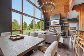 Un vaste espace de vie doté d’une grande table en bois, de sièges confortables, d’une cheminée en pierre et offrant une vue sur l’extérieur.