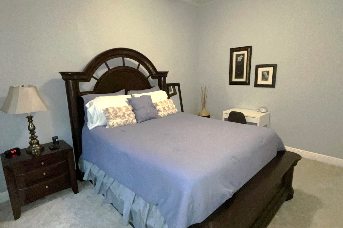 A bedroom with a bed, nightstand, lamp, ceiling fan, and framed pictures on the wall.