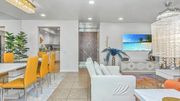 A modern living room with a white sofa, yellow chairs, a dining area with a white table, and a TV displaying a beach scene.