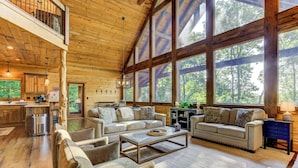 A spacious cabin with a high ceiling, large windows, a cozy living area with a sofa and chairs, a kitchen with wooden cabinets, and a view of the surrounding forest. - Hot Tub Soaks, Scenic Vistas, Starlit S'mores, Game Nights & Outdoor Fun - Little Creek Cabin (Blue Ridge)
