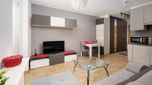 A modern living room with a flat-screen TV, a glass coffee table, and a sofa. There is a dining area with a red table and chairs. The kitchen is visible with white cabinets and a microwave. - Włochy Popularna Charming Studio (Warszawa)