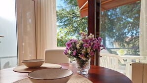 Ein Esstisch mit einer Schüssel und einem Blumenstrauß, ein Stuhl sowie Blick auf Bäume durch ein Fenster.