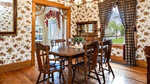 A dining room with a wooden table and chairs, floral wallpaper, a mirror, and a window with curtains.