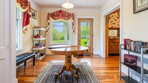 A room with a wooden floor, a round table with a bench, a bookshelf, and a door leading to a garden.