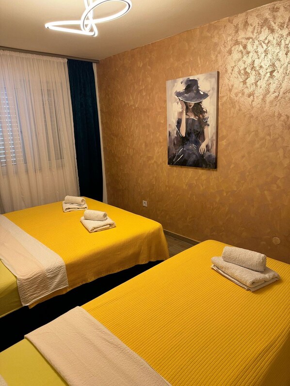 A hotel room with two beds, each with a yellow bedspread and white towels. There is a painting on the wall and a ceiling light fixture.