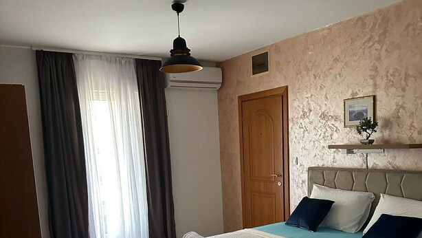 Egy hálószoba, amelyben van egy ágy, egy fa ajtó, egy mennyezeti ventilátor és egy függönnyel ellátott ablak.