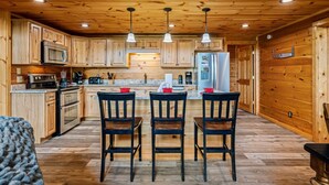 A wooden cabin interior with a kitchen featuring wooden cabinets, a microwave, stove, and refrigerator. There are three wooden chairs at a table with a rug.