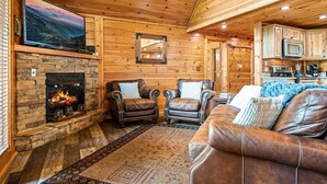 A cozy cabin interior with a stone fireplace, leather armchairs, a brown leather sofa, a mounted television, and a kitchen area.
