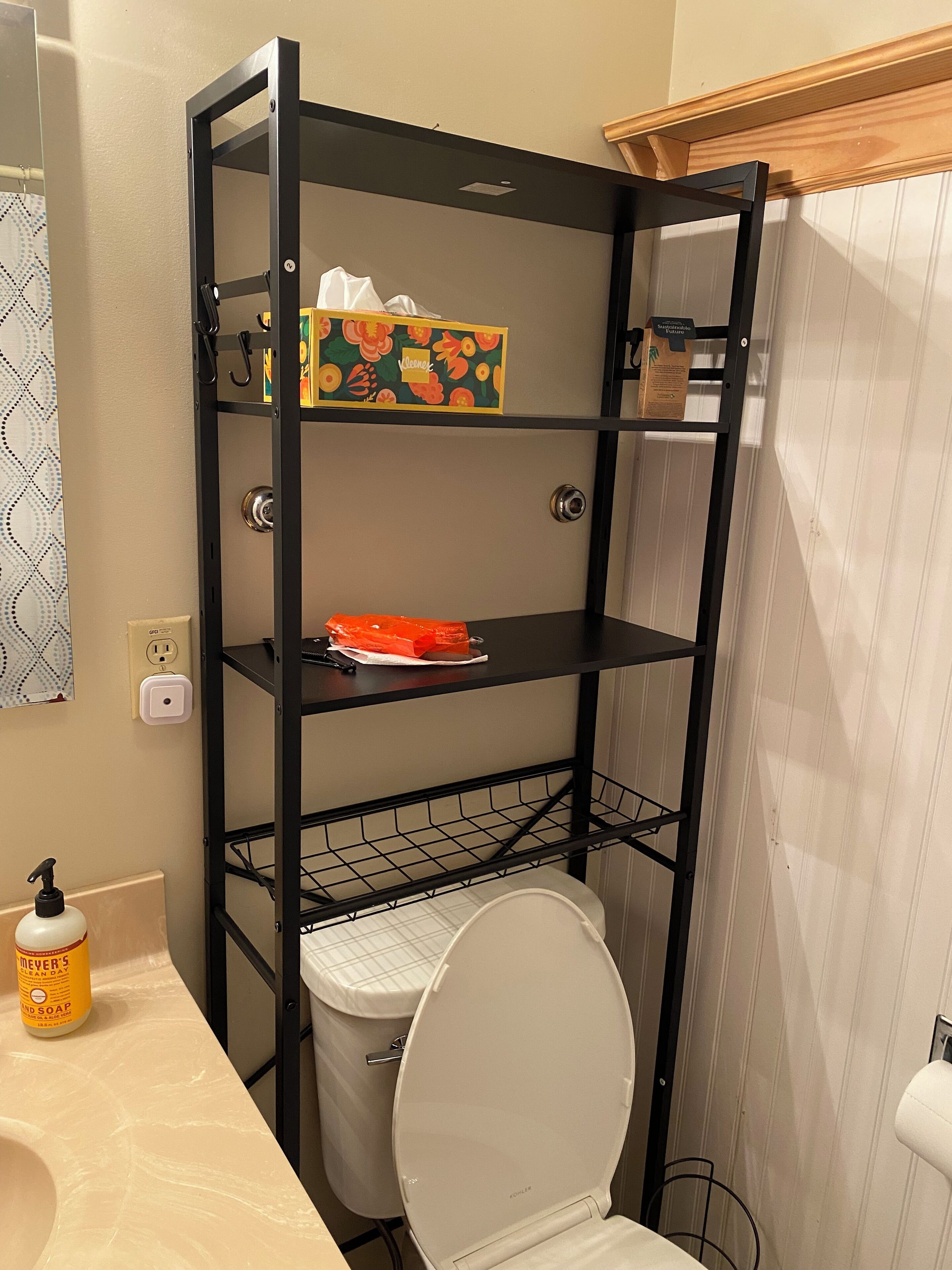 A bathroom with a black metal shelf unit, a toilet, a sink with a bottle of hand soap, and a shower curtain.