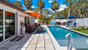 A pool area with a lounge chair and umbrella, a sign reading "BEACH HOUSE", and a pool with a tiled deck. - Fancy That: Pool, Putting Green, 1.5 Blocks to Beach by Anna Maria Life Vacation Rentals (Holmes Beach)