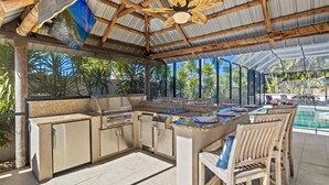 An outdoor kitchen with a grill, sink, and bar area, overlooking a pool and lush greenery.