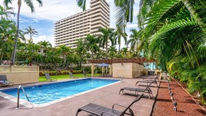 A swimming pool with lounge chairs, surrounded by palm trees and a large hotel building.