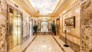 A luxurious hotel lobby with marble floors, gold accents, a chandelier, and potted plants.